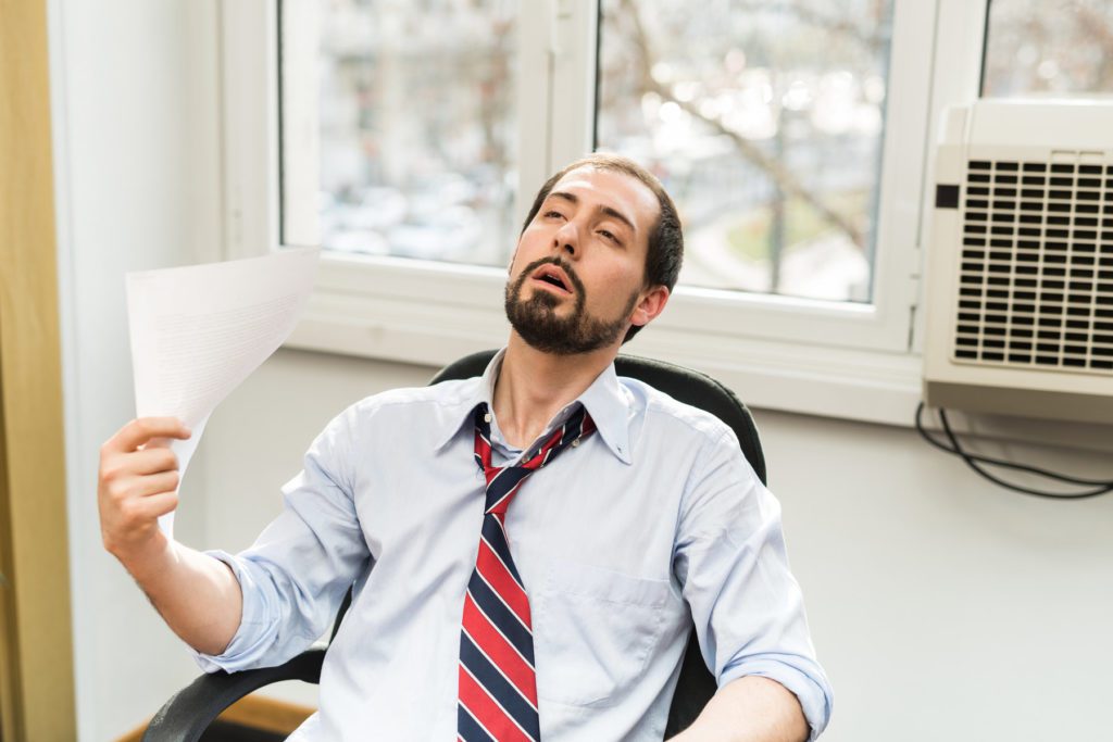 An overheated man fanning himself with paper in front of a non-functioning window a/c unit.