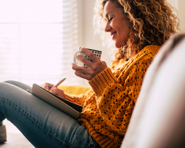 A woman writing into a journal in her lap while holding a mug in a reclined position.