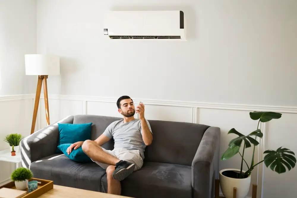 A man sitting in his living room under a mini split system, holding a remote.