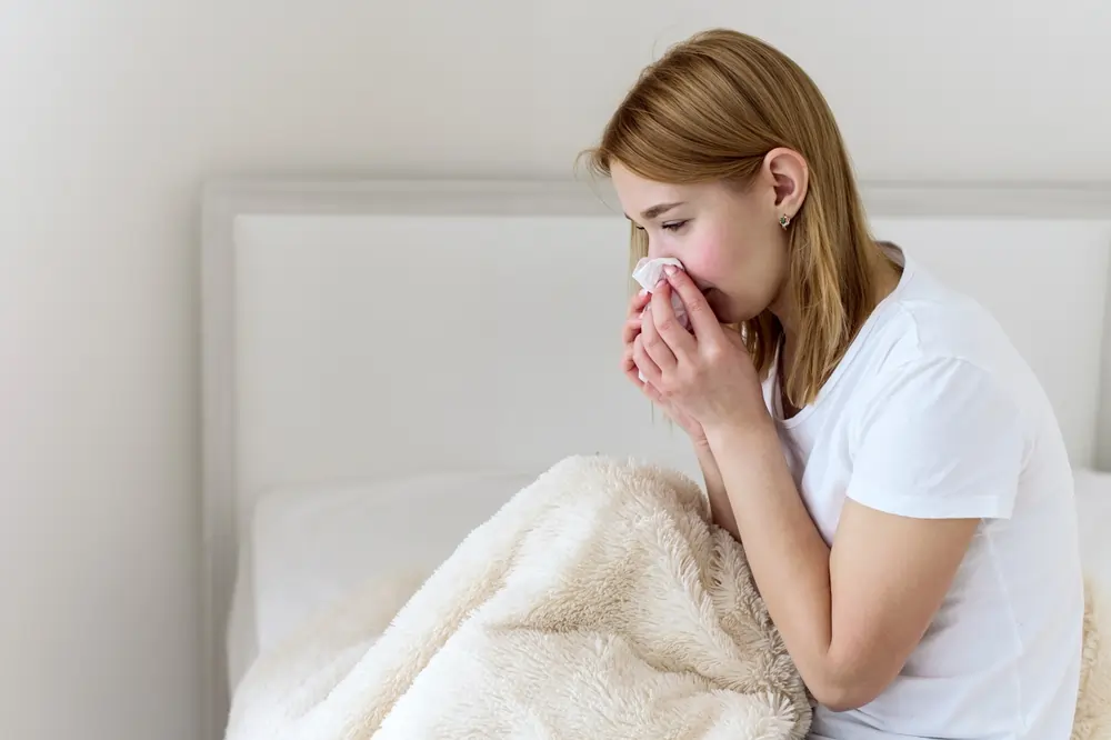 A young woman blowing her nose while sitting at the edge of her bed.