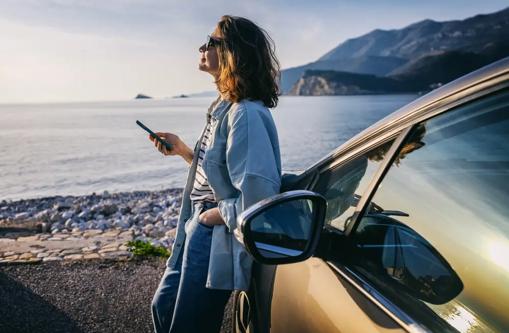 A woman in sunglasses looking at the sky while leaning against a car, phone in hand.